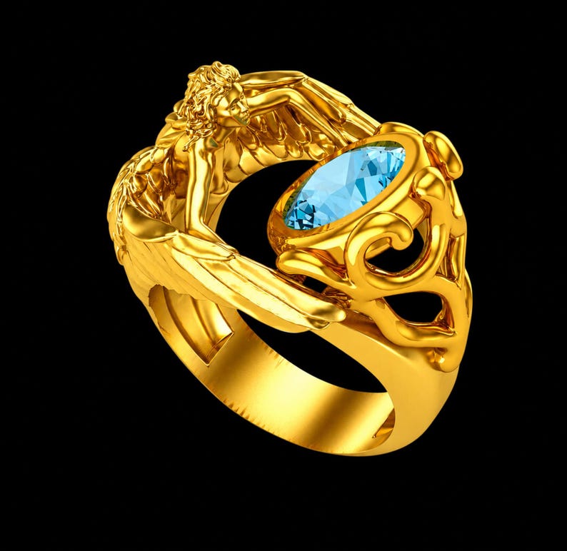May include: A gold ring featuring a sculpted figure with wings that wrap around the band. The ring is set with a single, oval-cut, light blue gemstone held within a decorative, swirling gold setting that connects to the winged figure's form.