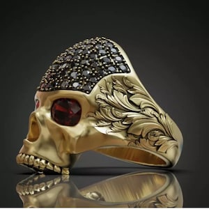 Puede incluir: Un anillo de calavera dorado con un diseño detallado. La frente de la calavera está cubierta de pequeñas piedras negras, y las cuencas de los ojos presentan gemas rojas. El anillo tiene intrincados grabados florales en los laterales.