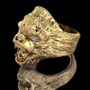 May include: A gold-toned ring featuring a detailed, three-dimensional gorilla head sculpture with its mouth open. The gorilla has textured fur detailing and two small, clear, round stones set into the eye sockets, reflecting light.