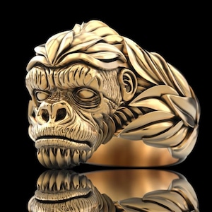 May include: A gold-toned ring featuring a highly detailed, three-dimensional sculpted gorilla head as the centerpiece. The band of the ring is intricately designed with a pattern of overlapping, textured leaves that wrap around the finger.