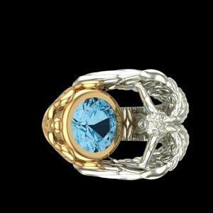 May include: A ring featuring a round, light blue gemstone set in a gold bezel. The band is crafted from silver-toned metal and shaped like two symmetrical wings that meet at the centre, accented with a small, clear, round-cut stone at the junction.