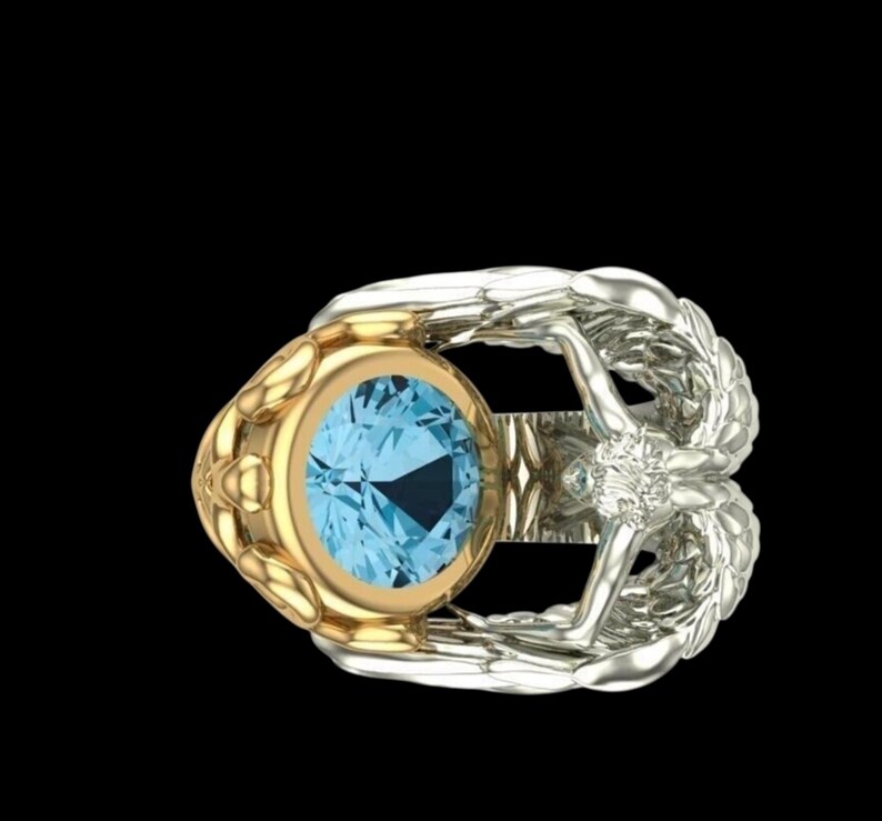 May include: A ring featuring a round, light blue gemstone set in a gold bezel. The band is crafted from silver-toned metal and shaped like two symmetrical wings that meet at the centre, accented with a small, clear, round-cut stone at the junction.