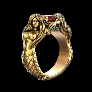 May include: A gold-colored ring featuring two mermaid figures. The mermaids have flowing hair and are adorned with small blue and red gemstones. The ring's design is intricate, with a focus on the mermaids' forms and the scales of their tails.