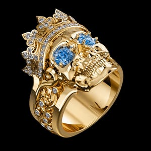 May include: A gold-colored ring featuring a skull design with a crown and blue gemstone eyes. The ring is adorned with small, clear gemstones and intricate detailing. The ring is set against a black background.