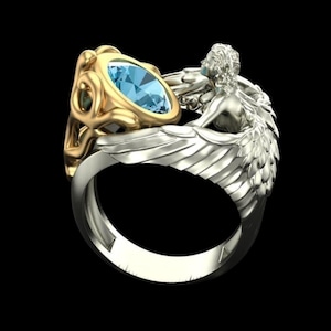 May include: A two-tone ring featuring a central oval-cut light blue gemstone set in a gold-coloured frame. The band is silver-coloured and incorporates a sculpted figure with detailed wings that wrap around the side of the gemstone setting.