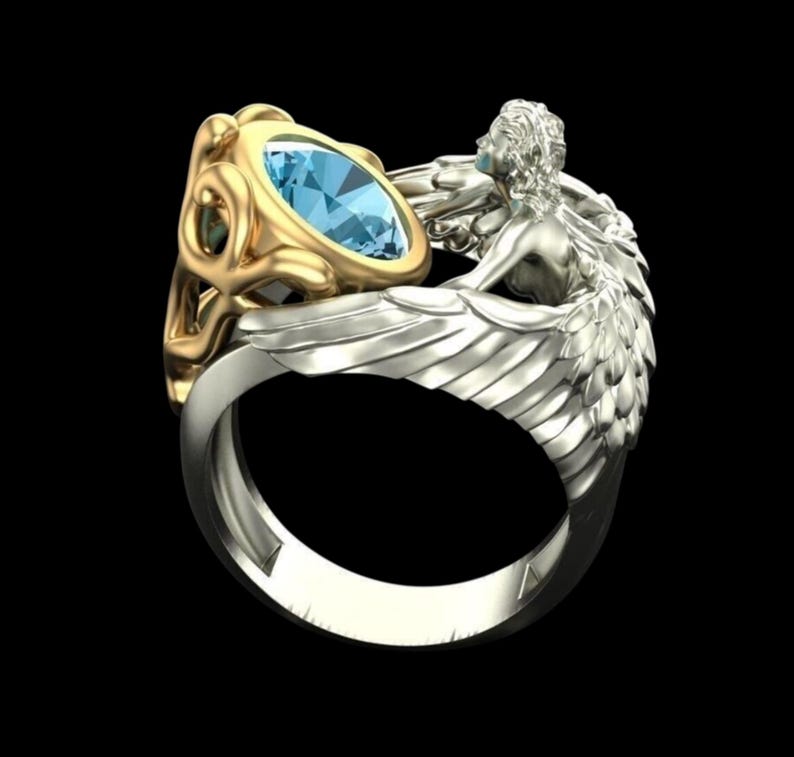 May include: A two-tone ring featuring a central oval-cut light blue gemstone set in a gold-coloured frame. The band is silver-coloured and incorporates a sculpted figure with detailed wings that wrap around the side of the gemstone setting.