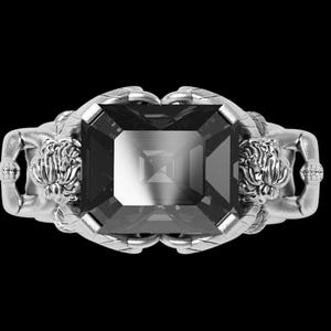 Peut inclure: Bague en argent avec une grosse pierre pr&eacute;cieuse rectangulaire gris fonc&eacute;. La monture de la bague pr&eacute;sente deux figures sculpt&eacute;es tenant la pierre pr&eacute;cieuse. Les figures ont des cheveux d&eacute;taill&eacute;s et sont positionn&eacute;es de chaque c&ocirc;t&eacute; de la pierre.