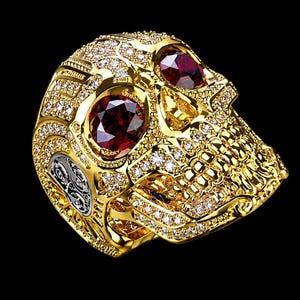 May include: A gold ring shaped like a skull, featuring two large, round, deep red gemstones set in the eye sockets. The entire surface of the skull is encrusted with numerous small, clear, sparkling stones, and the side displays intricate silver-toned metal detailing.