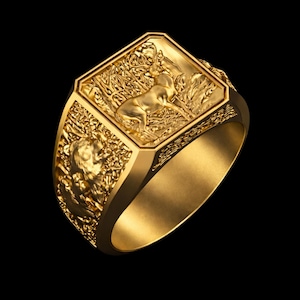 Peut inclure: Une bague dorée avec un design détaillé. La bague présente une face rectangulaire avec un relief d'un cerf dans une forêt. La bande a des détails en relief supplémentaires. La bague est sur fond noir.