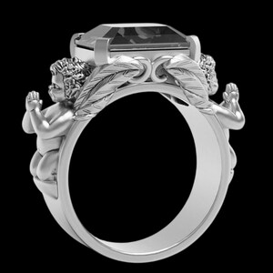 Peut inclure: Bague en argent orn&eacute;e d'une grosse pierre rectangulaire. Deux ch&eacute;rubins encadrent la gemme, les mains jointes en pri&egrave;re. Le design de la bague incorpore des d&eacute;tails en forme de plumes, cr&eacute;ant un bijou orn&eacute; et d&eacute;taill&eacute;.