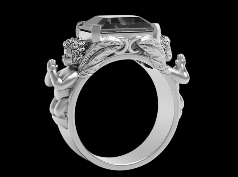 Peut inclure: Bague en argent orn&eacute;e d'une grosse pierre rectangulaire. Deux ch&eacute;rubins encadrent la gemme, les mains jointes en pri&egrave;re. Le design de la bague incorpore des d&eacute;tails en forme de plumes, cr&eacute;ant un bijou orn&eacute; et d&eacute;taill&eacute;.