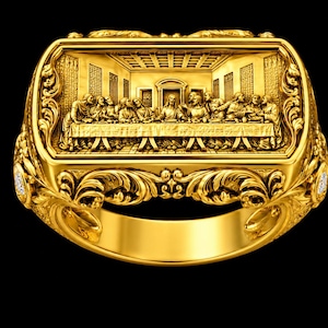May include: A gold ring featuring a rectangular face with a detailed relief carving of The Last Supper. The band and sides of the ring are decorated with intricate scrollwork and filigree patterns, with small embedded stones on the sides.