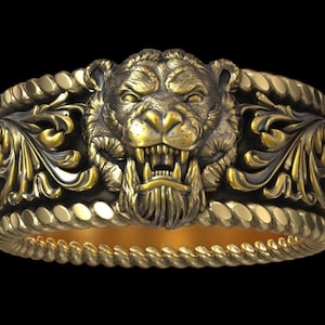 May include: A gold-colored ring with a detailed lion's head design. The ring features intricate floral and rope-like patterns. The lion's face is centered, with a fierce expression. The ring is set against a black background.