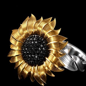 May include: A gold-coloured sunflower ring with a black centre. The ring's band is silver, and the sunflower petals are detailed. The ring is set against a black background.