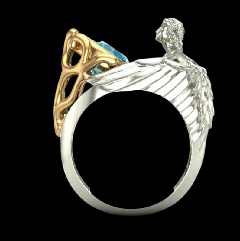 May include: A ring featuring a silver-toned band shaped like a pair of wings with a sculpted figure at the top. A gold-toned setting holds a light blue gemstone, with intricate metalwork detailing the side of the band opposite the wings.