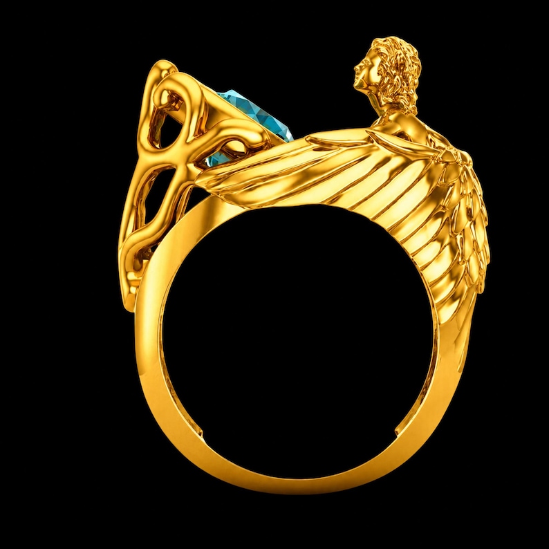 May include: A gold ring featuring a sculpted figure with detailed wings extending along the band. The ring is set with a single, faceted, light blue gemstone held in a decorative, openwork gold setting that rises above the band's circular frame.