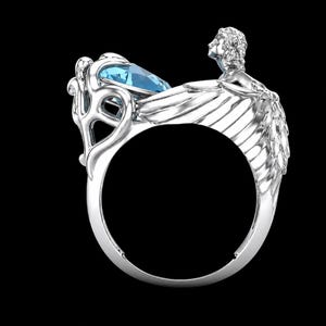 May include: A silver ring featuring a faceted light blue gemstone set within a decorative frame. The band is shaped like a pair of feathered wings extending from a sculpted figure of a person with flowing hair, positioned above the gemstone setting.