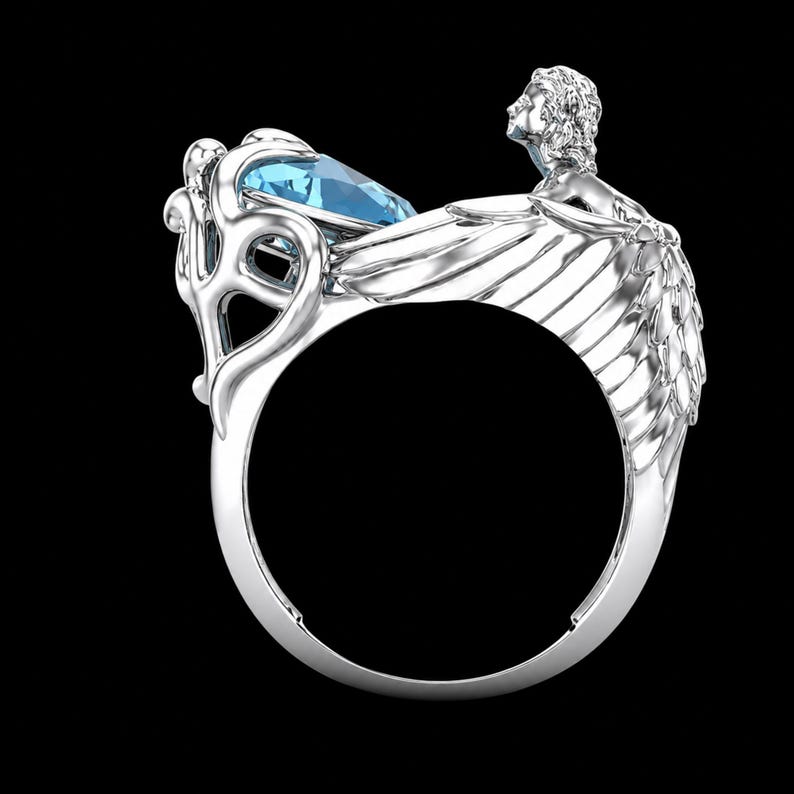 May include: A silver ring featuring a faceted light blue gemstone set within a decorative frame. The band is shaped like a pair of feathered wings extending from a sculpted figure of a person with flowing hair, positioned above the gemstone setting.