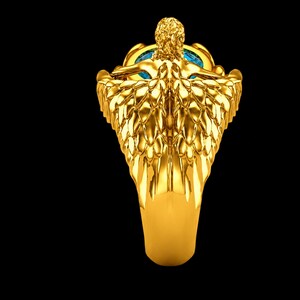 May include: A gold ring featuring a sculpted figure with detailed wings extending down the band. The figure is positioned above a round, bright blue gemstone set into the centre of the ring, creating a prominent focal point with metallic texture.