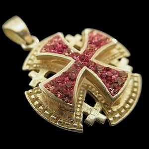 May include: A gold-colored pendant with a cross design. The cross is filled with small, pink gemstones. The pendant has a loop at the top for a chain and is set against a black background.