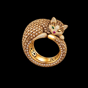 May include: A gold ring shaped like a curled cat, featuring a textured mesh body encrusted with numerous small clear diamonds. The cat has a single green gemstone eye, a small pink gemstone nose, and diamond-paved ears and face details.