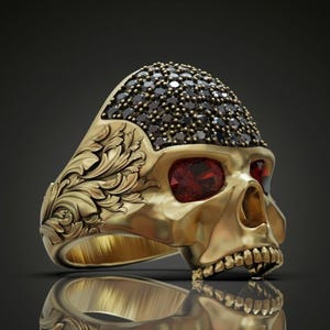 May include: A gold-colored skull ring with a detailed design. The skull's forehead is covered in small black stones, and the eyes are red. The ring has a detailed floral pattern on the side.