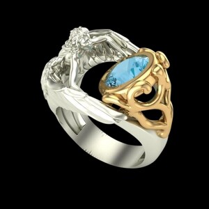 May include: A two-tone ring featuring a silver-coloured band shaped like a winged figure with detailed feathers. The ring holds a central oval-cut light blue gemstone set within a decorative gold-coloured metal frame with intricate scrollwork.