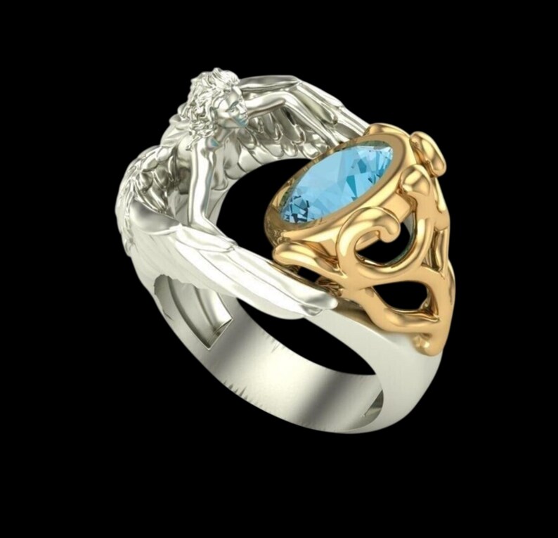 May include: A two-tone ring featuring a silver-coloured band shaped like a winged figure with detailed feathers. The ring holds a central oval-cut light blue gemstone set within a decorative gold-coloured metal frame with intricate scrollwork.