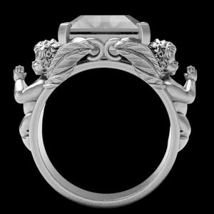 Peut inclure: Une bague en argent orn&eacute;e d'une grosse pierre pr&eacute;cieuse rectangulaire. La conception de la bague comprend deux ch&eacute;rubins de chaque c&ocirc;t&eacute; de la pierre pr&eacute;cieuse, avec des accents de plumes d&eacute;taill&eacute;s. La bague est pos&eacute;e sur un fond noir.