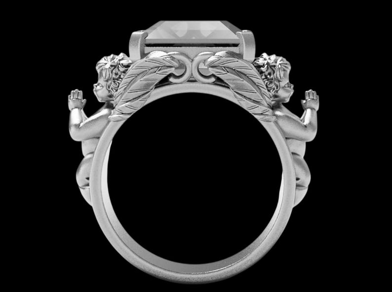 Peut inclure: Une bague en argent orn&eacute;e d'une grosse pierre pr&eacute;cieuse rectangulaire. La conception de la bague comprend deux ch&eacute;rubins de chaque c&ocirc;t&eacute; de la pierre pr&eacute;cieuse, avec des accents de plumes d&eacute;taill&eacute;s. La bague est pos&eacute;e sur un fond noir.