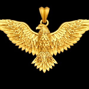May include: A gold-colored pendant in the shape of an eagle with outstretched wings. The eagle has detailed feathering and a loop at the top for a chain. The background is solid black.