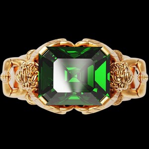 Peut inclure: Une bague en or avec une grosse pierre verte taill&eacute;e en &eacute;meraude. La bague pr&eacute;sente des d&eacute;tails dor&eacute;s orn&eacute;s, notamment des figures sculpt&eacute;es de chaque c&ocirc;t&eacute; de la pierre. Le fond est noir.
