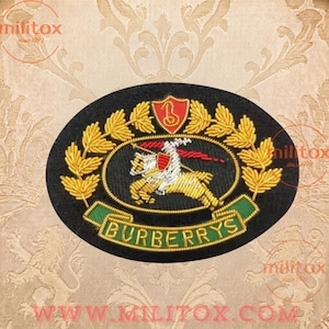Op de afbeelding: Een ovale zwarte stoffen patch met een geborduurde gouden ridder te paard, een rood schild met een gouden letter B, gouden laurierbladeren en een groene banner met de gouden tekst BURBERRYS. Het ontwerp is gedetailleerd met metaaldraad.