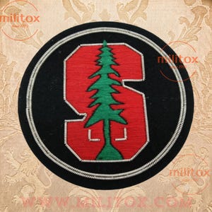 May include: A circular embroidered patch featuring a black background with a large red letter S outlined in silver thread. A green pine tree is stitched vertically through the center of the letter. The entire patch is bordered by a silver corded edge.