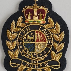 Pode incluir: Um emblema de tecido preto com bordado metálico dourado apresentando um brasão com uma coroa no topo. O emblema circular central exibe o texto RALPH LAUREN, um leão e letras de monograma. Uma faixa na parte inferior diz LAUREN em fio dourado.