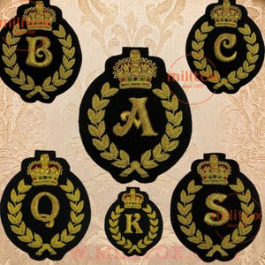 May include: Six black circular embroidered patches featuring gold thread laurel wreaths surrounding a central letter. Each patch is topped with a detailed gold, red, and green crown. The letters A, B, C, K, Q, and S are displayed in gold thread.