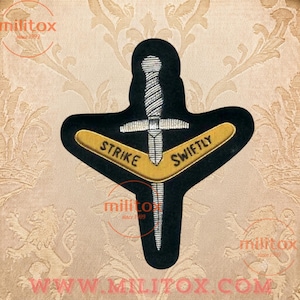 May include: A black embroidered patch featuring a silver dagger positioned vertically behind a gold-coloured boomerang shape. The text STRIKE SWIFTLY is stitched in black capital letters across the gold boomerang. The patch has a distinct shape.