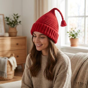 Può includere: Un berretto in maglia rosso brillante con un motivo a nido d'ape, un ampio risvolto a coste e una parte superiore lunga e appuntita che si curva verso il basso, terminando con una piccola nappa di filo pendente. Il berretto è in filato spesso.