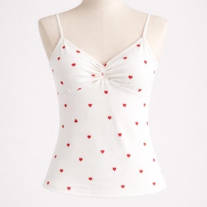 Op de afbeelding: Witte camisole top met een patroon van rode hartjes. De top heeft dunne spaghettibandjes en een geplooid detail bij de buste. De stof is een zacht, lichtgewicht materiaal.