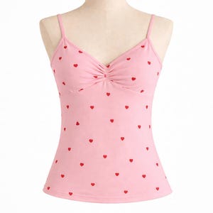 Op de afbeelding: Een lichtroze camisole top met kleine rode hartmotieven. De top heeft fijne spaghettibandjes en een geplooid detail bij de buste. De stof voelt zacht aan, met harten gelijkmatig verdeeld.