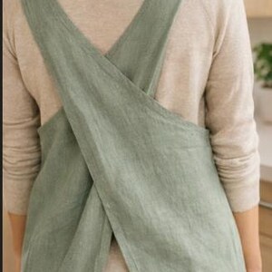 May include: A sage green linen apron with crisscross straps. The apron is made of a soft, textured fabric and has a simple, minimalist design. The apron is worn over a light-colored long-sleeved shirt and dark jeans.