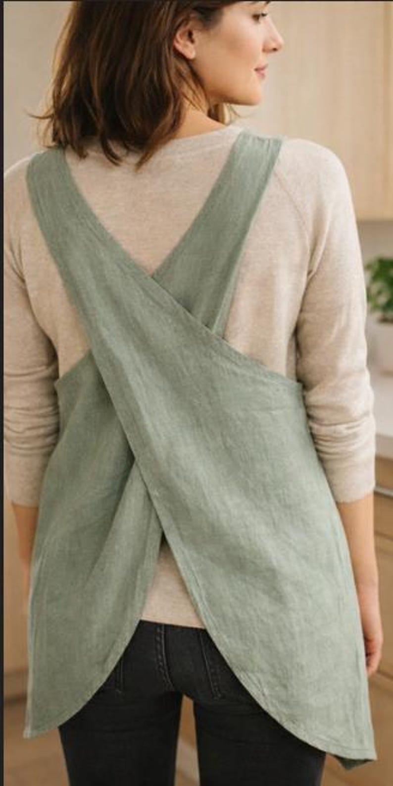 May include: A sage green linen apron with crisscross straps. The apron is made of a soft, textured fabric and has a simple, minimalist design. The apron is worn over a light-colored long-sleeved shirt and dark jeans.