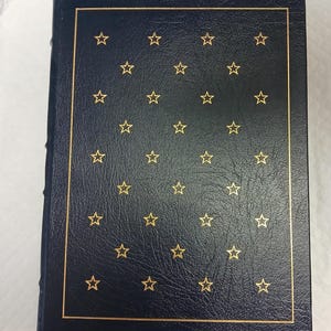 May include: A navy blue book with a gold border and gold star pattern. The cover has a textured appearance. The spine is visible on the left side.