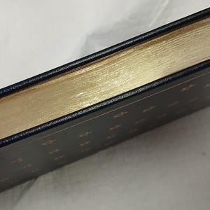 May include: A hardcover book with gold gilded edges and a dark blue cover. The edges of the pages are a bright gold color, and the cover has gold lettering. The book is bound in black leather.
