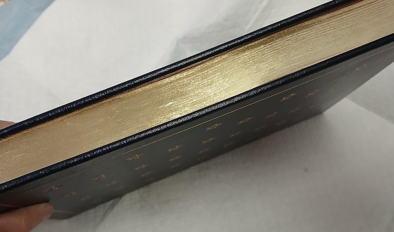 May include: A hardcover book with gold gilded edges and a dark blue cover. The edges of the pages are a bright gold color, and the cover has gold lettering. The book is bound in black leather.