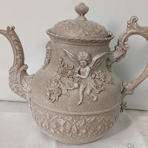 Op de afbeelding: Beige keramische theepot met een gedetailleerd reliëf ontwerp. De theepot heeft een engel, rozen en sierlijke krullen. Het heeft een gebogen tuit, een decoratief handvat en een deksel met een knop. De theepot is een decoratief item.