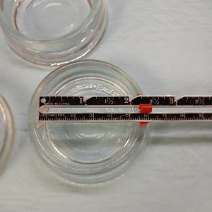 May include: Clear glass votive candle holders, approximately 5 cm in diameter, are shown with a ruler. The votives have a simple, round design with a slightly raised base. The image is taken from above.