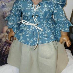 May include: A doll wearing a blue floral jacket with a hood and ruffled sleeves, laced with a white cord. The jacket is paired with a beige skirt. The outfit is designed for a doll.