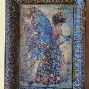 May include: A framed artwork featuring a fairy with blue wings and a floral dress, holding a butterfly. The artwork is set within a decorative, ornate, dark-toned frame with a stand.