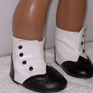 May include: A pair of doll shoes with a classic design. The shoes are two-tone, featuring white uppers and black toes. They have button closures and are designed to resemble vintage footwear.